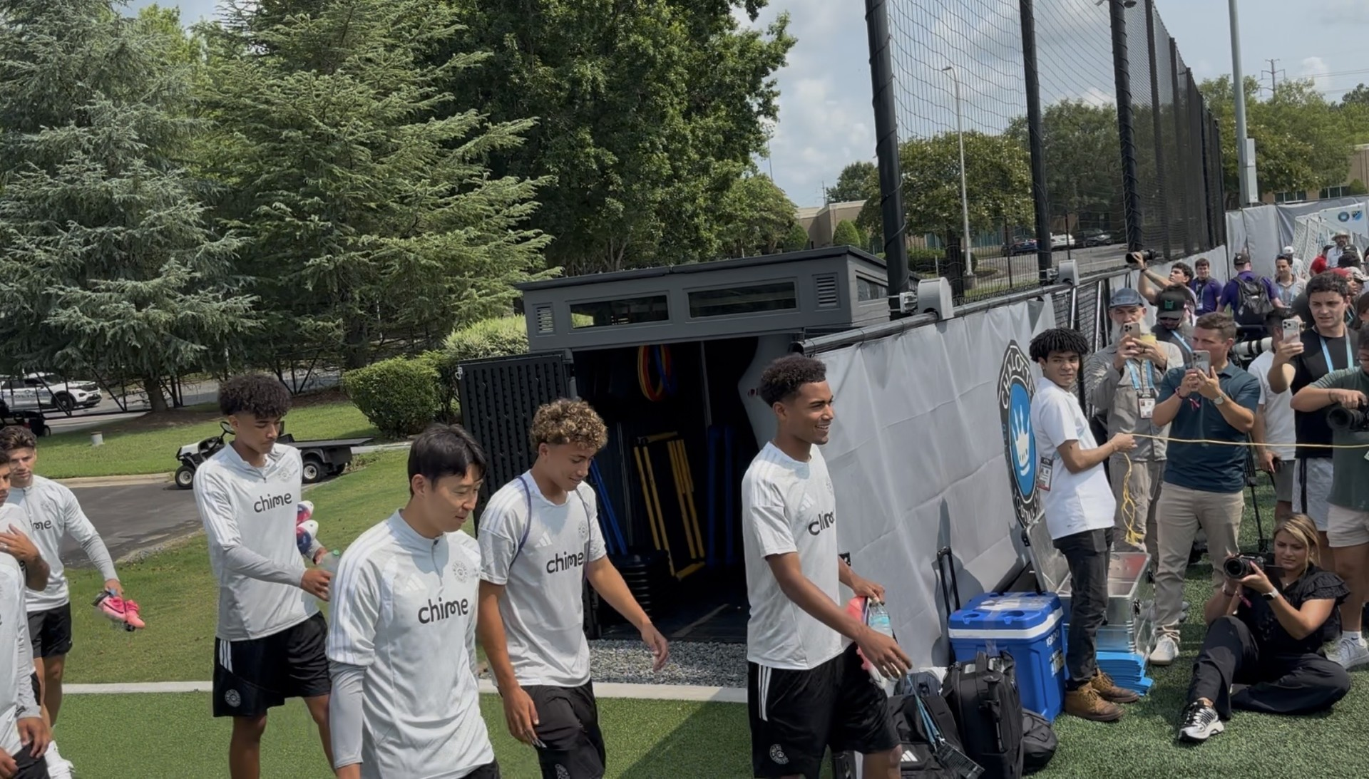 A group of young people wearing white shirts with the "Chime" logo standing in front of a large black trailer on a grassy field, with spectators watching in the background.